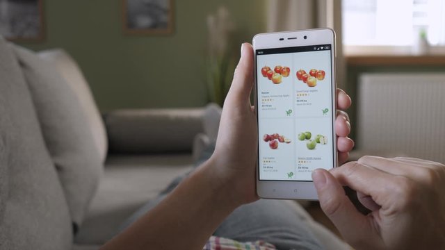 Woman Orders Food Home In An Online Store Using a Smartphone. Female Selects Fruit Apples in Grocery Online Store. Woman at Home Lying on Couch in Living Room Using Smartphone Buys in Internet Shop.