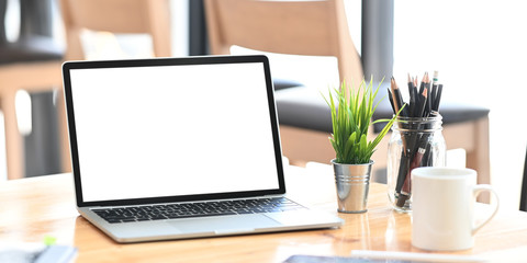 Fototapeta premium Blank screen computer laptop putting on wooden working desk and surrounded by potted plant, pencil holder and ceramic coffee cup. Orderly workspace and space for advertisement concept.