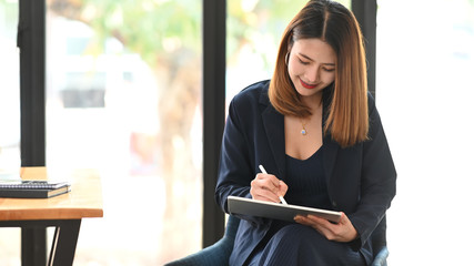 Photo of sexy businesswoman holding a computer tablet and stylus pen while sitting at the wooden working desk over comfortable sitting room as background.