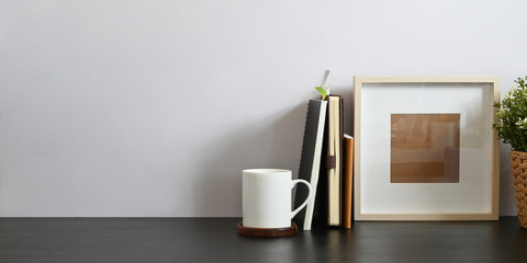 Working desk along with book, notebook, picture frame, potted plant and coffee cup putting together on it with white cement wall as background. Orderly workspace concept.