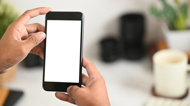 Cropped image of smart man's hands holding a cropped black smartphone with white blank screen over his white working desk as background.