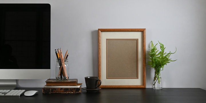 Photo of black working desk along with black blank screen computer monitor, books, notebook, pencil holder, picture frame, potted plant putting together on it with white cement wall as background.