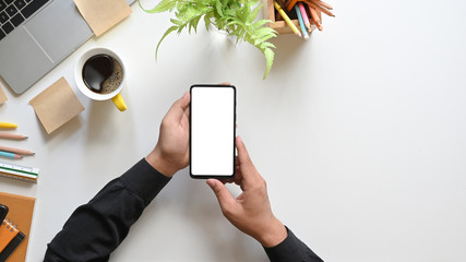 Top view hands holding a white blank screen smartphone over coffee cup, potted plant, pencils,...