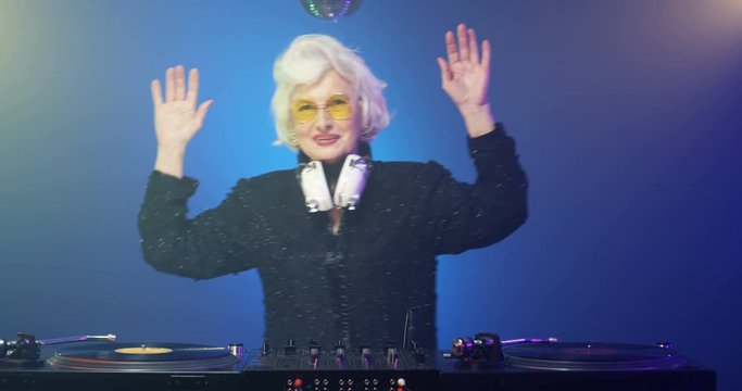 Retro style funny Caucasian old granny mixing music at party. Clubbing grandma DJ with headphones dancing while playing disco on dj mixer. Vintage grey-haired lady partying at club. 70s concept