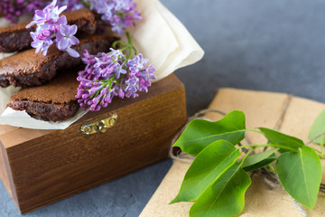 Romantic still life with lilac flowers and brownie, wet cake. Dessert for served for tea or coffee break in wooden box. Snack on a spring day in the garden.
