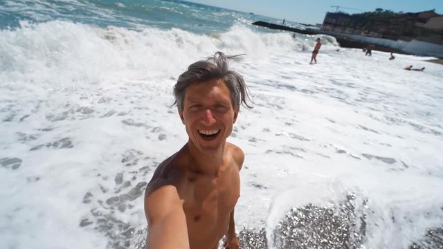 happy laughing tanned athletic young man taking video selfie while walking along beach with waving sea at summer vacation resort. slow motion