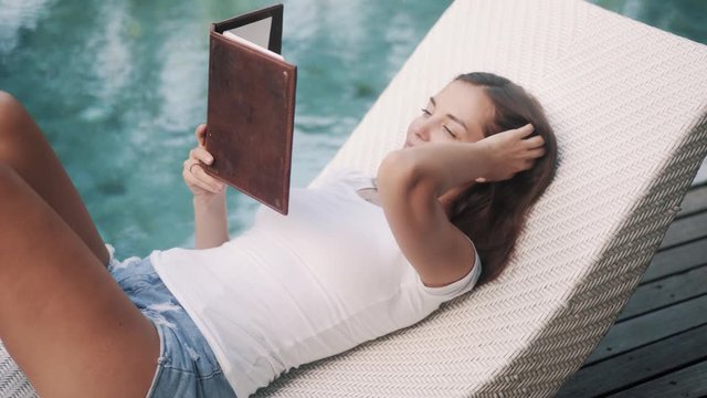 Portrait of woman in summer clothes lies on deck chair and reads book near pool