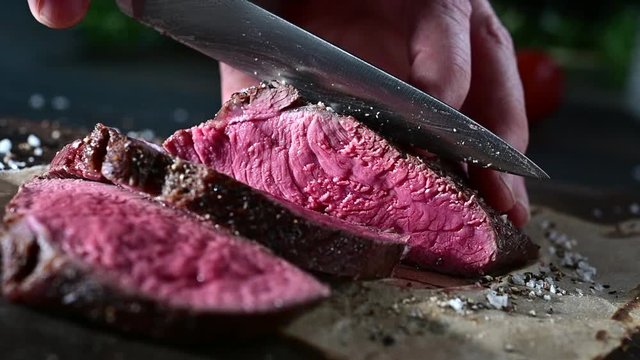 Man slicing through a portion of seasoned rare roast beef steak with a large carving knife in a close up on the meat and the blade