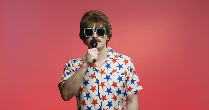 Joyful male model over dark pink background. Funny retro style dark-haired Caucasian man with mustache singing in microphone. Vintage singer guy in shirt with stars posing in studio. 50s concept