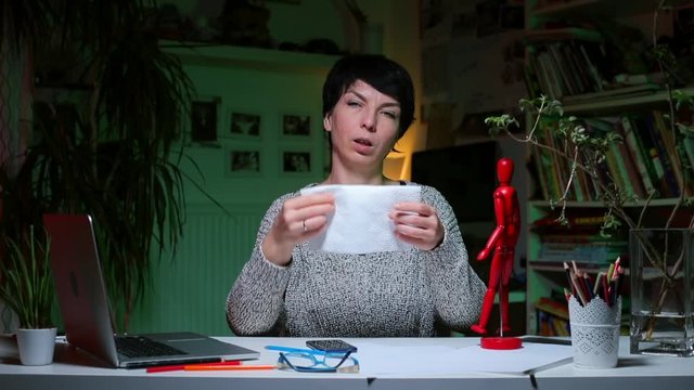 A woman at her home office table blows her nose in a napkin. Cold in the workplace. Removing sputum from the nose.