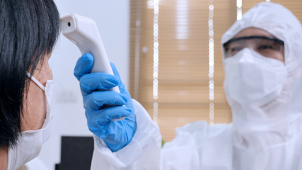 Asian doctor in protective PPE suit using infrared thermometer for checking health of virus...