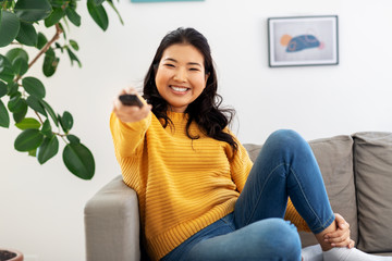 people and leisure concept - happy smiling asian young woman with tv remote control sitting on sofa at home