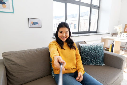 people and leisure concept - happy smiling asian young woman in yellow sweater taking picture with selfie stick at home