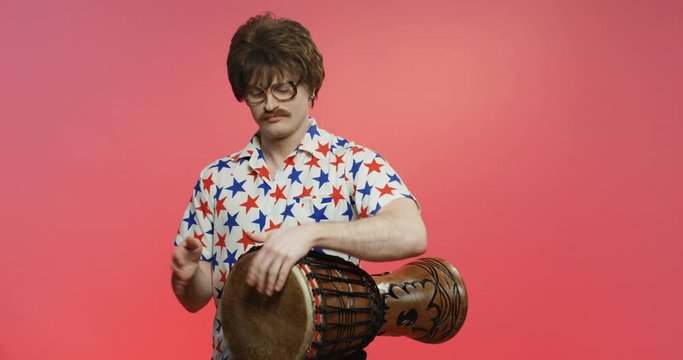 Stylish male model with eyeglasses in shirt with stars playing on vintage musical instrument. Funny Caucasian man with smile on face over dark pink background with Doumbek. Retro style concept