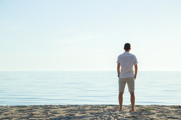 Young man in white clothes standing alone on sand and staring at calm sea and light blue sky. Fresh air. Empty place for emotional, sentimental, inspirational text, quote or sayings. Back view. - Powered by Adobe