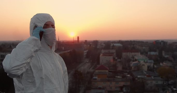 Portrait of a young male in protective suit and a respiratory mask standing and turning around and answering a phone call on a roof with a cityscape view in a sunny spring sunset during pandemic