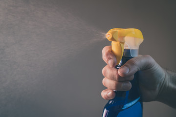 Bare hand holding a spray bottle for cleaning and operating it. Drops from bottle are being sprayed all over the place. Frontal view of an operational plastic spray bottle.