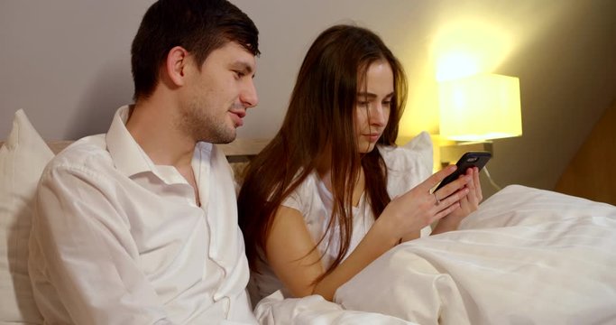 woman is watching social nets by cell phone in bed before sleep, her husband is sitting and talking