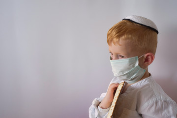 A boy in a kippah and a medical mask holds a Passover matzo in his hands. Free space for text.
