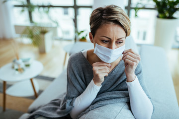 Pensive woman feeling worried and sitting at home with protective mask on her face during virus...