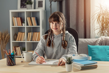 Smiling teen girl sitting at desk looking at camera, studying from home, front view