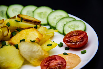 Homemade fried potatoes with fresh cucumbers, cherry tomatoes and sauces on the white plate. Delicious ukrainian fried potatoes with vegetables against background. Cooked tasty meal for lunch.