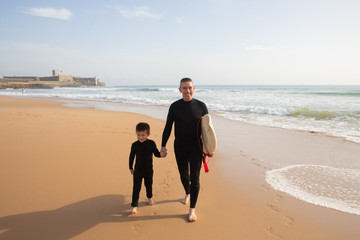 Smiling father holding son hand and carrying surfboard. Sea on background. Man and boy in full body swimsuits leaving footprints on sand. Vacation, surfing and summer concept