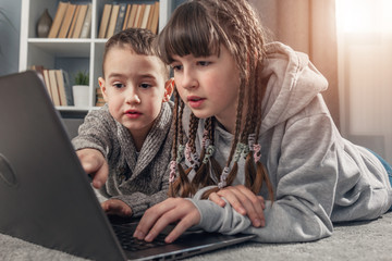 2 kids, girl and boy, browsing internet on laptop lying on floor at home during quarantine