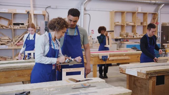 Dolly shot of female tutor with carpentry student in workshop studying for apprenticeship at college using wood plane - shot in slow motion