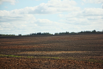 A vast brown ploughed field hill against a cloudy blue sky and trees on horizon on a spring day — land cultivation, agriculture, rural landscape