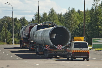 Long oversized cargo transportation logistics, large long tubes on low-frame trawls trucks with an escort car on asphalt road