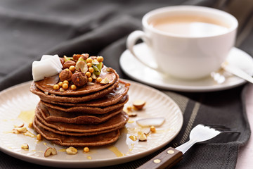 chocolate pancakes with kiwi, hazelnut, honey, coconut flakes on plate with fork and cup of tea or coffee on grey towel background