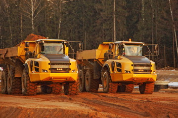 Two yellow heavy dump trucks loaded and empty on a construction site against the background of a forest, front view
