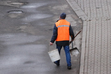 A janitor man in an orange vest walks with a bucket and scoop along an asphalt street, top view...