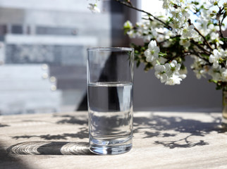 Transparent glass with clear water on a background of flowering branches. The concept of health and beauty. Side view.