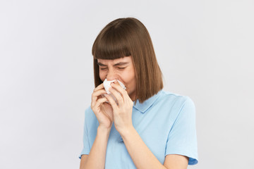 female with dark hair and in blue shirt blows nose with eyes closed on grey background