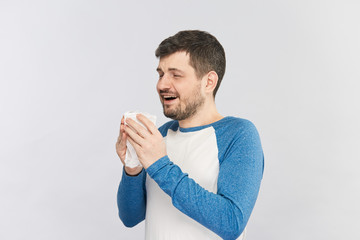happy young bearded man start to sneeze into his white tissue isolated on grey background
