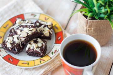Closed-up of homemade chocolate brownies with almond nuts and white chocolate chips topping served with coffee on white wooden background.