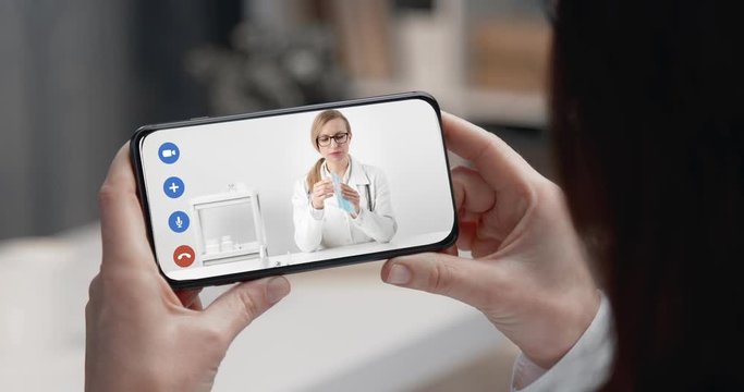 View from back of mature woman sitting at home and watching video instruction on smartphone about how properly wearing protective mask. Protection from virus and advice from competent doctor.