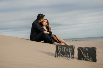 Pareja enamorados sentados en la arena vestidos de negro , con el mar y cielo de fondo. Save the date borroso.