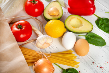 vegetables and fruit spilled from a paper grocery bag on a white wooden background, avocado,  tomatoes, peppers, lettuce, pasta, garlic, lemon,  onions, spinach, eggs, detox;  rustic; bio;