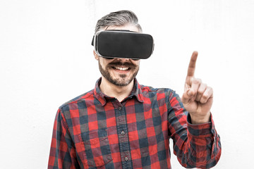 Content man in VR headset pointing with finger. Front view of cheerful bearded man in checkered shirt using virtual reality headset on grey background. Technology concept