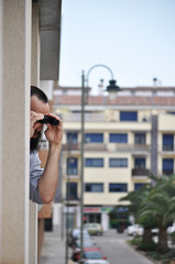 Young man leaning out of apartment window looking through binoculars in a Spanish Village in quarantine time. Man watching condominiums with binoculars