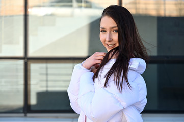 Outdoors portrait of a fashionable young brunette girl in a white jacket stands against the...