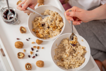 Happy mom and daughter have healthy breakfast in bed on a sunny morning. Oatmeal porridge with nuts and raisins. Hands close up. Healthy food concept