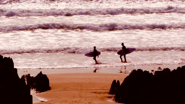 Two unrecognizable people in silhouette, walking in the surf along a rocky beach carrying surfboards, as multiple small waves break on the sand. Woolacombe, Devon, England, UK.