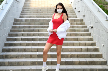 Outdoor portrait of a fashionable young brunette girl in a medical mask in a red dress and a white jacket stands on the steps of the stairs.