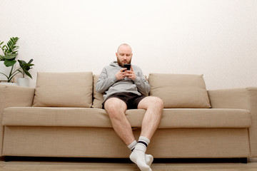 A young man with a beard in home clothes is sitting on a sofa and is using a smartphone. Smartphone...