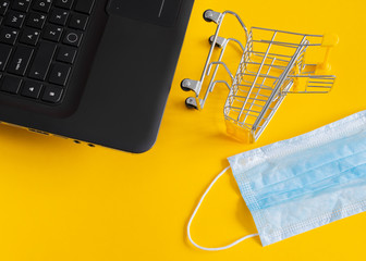 Text space: an empty grocery cart lies on its side next to a black laptop and a protective mask on a yellow background during the quarantine period, top view. Delivery, online shopping