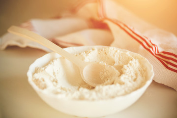On a white table is a white bowl of crumbly wheat flour, in which is a wooden elegant spoon, and next to the plate is a white towel with red stripes, illuminated by sunlight.